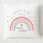 A smiling mother holding her newborn baby in a nursery setting. She is sitting in a white armchair next to a white personalised rainbow cushion that says Welcome to the WORLD with the baby's name and birth date.