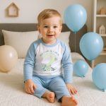 Toddler boy wearing personalised blue and white dinosaur birthday pyjamas that say When I wake up I will be 2 and a boys name , sitting on a bed with blue balloons.