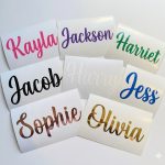 A flat lay arrangement of eight custom permanent vinyl name decals resting on their white backing paper against a clean white background, showcasing the names Kayla, Jackson, Harriet, Jacob, Harry, Jess, Sophie, and Olivia in a variety of glossy and metallic finishes including hot pink, purple, and rose gold.